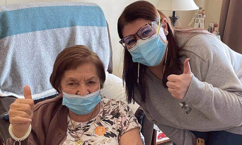 A senior and her caretaker wear masks and hold thumbs up to the camera.