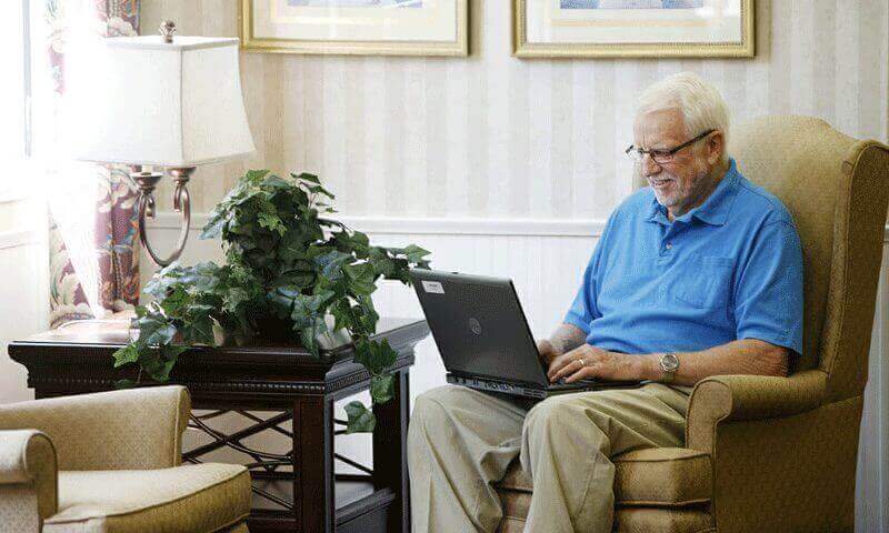 An elderly man uses a laptop in sitting in an armchair.